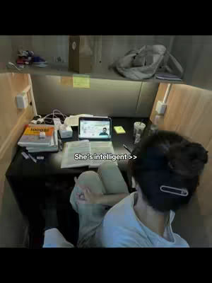 The image shows a desk in a study area with books, a laptop displaying a person's face, and a person's back facing the camera. The text overlay reads, "She's intelligent >>". The lighting and composition give a sense of focus and study.