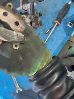 The image shows a close-up of a gloved hand working on a mechanical component. The hand, covered by a green glove, holds a metallic piece that appears to be part of a vehicle's braking system. A wrench and screwdriver are visible on a blue surface, indicating a repair or maintenance task.