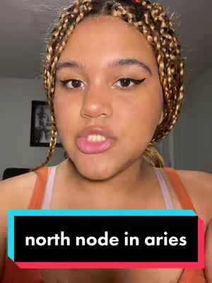 A close-up shot features a young woman with light brown skin, visible from the chest up. She has curly brown and blonde braids and is wearing a coral-colored top. Her eyebrows are arched, and she has eyeliner on, framing her dark eyes, and light pink lipstick. The background shows a room with a picture frame.