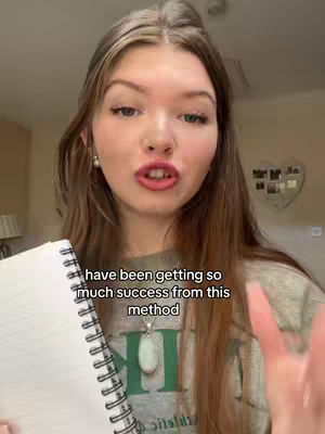 A young woman with long brown hair, wearing a green shirt, is holding a white notebook and speaking directly to the camera. The background is out of focus but appears to be a home interior with a heart decoration visible. Text overlayed on the image says: 'have been getting so much success from this method'.