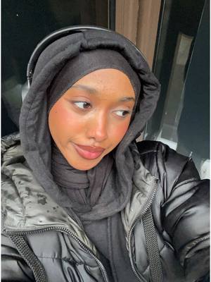 A young woman with brown skin wearing a black headscarf and a black hooded jacket stares to the side with a slight smile. Her cheeks have a pink blush, and she wears headphones. The background has a dark, blurred appearance.