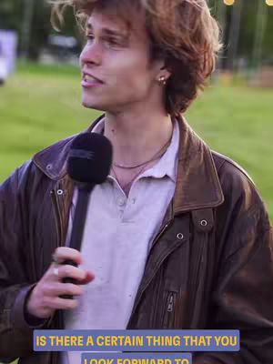 A young man with light brown hair and a leather jacket speaks into a microphone. The background shows a green, grassy area with out-of-focus details, possibly an outdoor event.