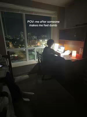 A night scene shows an interior room with a large window overlooking a cityscape lit up in the distance. A figure is seated at a desk, back to the camera, working on a laptop with a lamp on the desk. The room has a moody, low-light atmosphere.