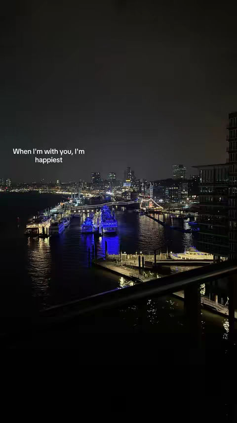 A nighttime city scene with a body of water in the foreground, boats and buildings in the midground, and city lights in the background. The sky is dark, and there are some clouds.