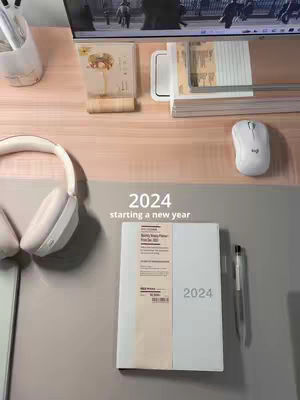 Top-down view of a desk setup with a variety of study-related items. A white notebook labeled '2024' lies on the desk, next to a pen. Overhead, a pair of white headphones sits on the desk. A computer screen displays what appears to be a cityscape. Above the notebook is the text '2024 starting a new year' in white font.
