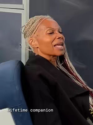 A woman with blonde braids is sitting in a chair, looking to her right, and slightly upward. The image is a close-up shot, and she is wearing a black coat or jacket. Below her is a caption that says, 'Lifetime companion'.