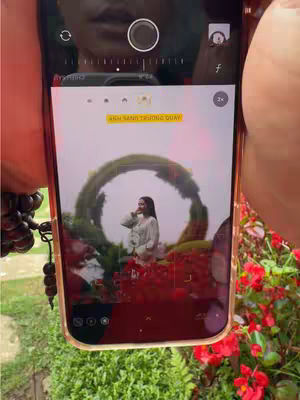 An image of a phone screen displaying a photograph of a woman posing in front of a floral arch at what appears to be a scenic location. The phone screen also shows the camera app interface. The phone is held up, and you can see hands holding the phone.
