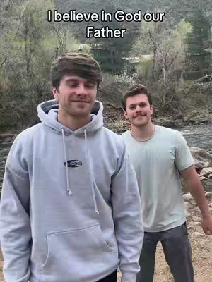Two young men stand outdoors near a river, one in a light gray hoodie with text above, and a second man in a light green t-shirt. The background features trees and the sky.