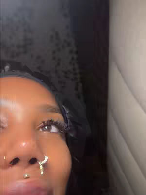A close-up shot of a person's eye and part of the nose, with multiple piercings, and a black head covering. The background is dark, with water droplets.