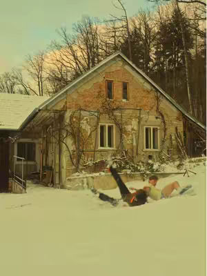 An abandoned house sits amidst a snowy landscape. Two individuals are sprawled in the snow in front of the building.
