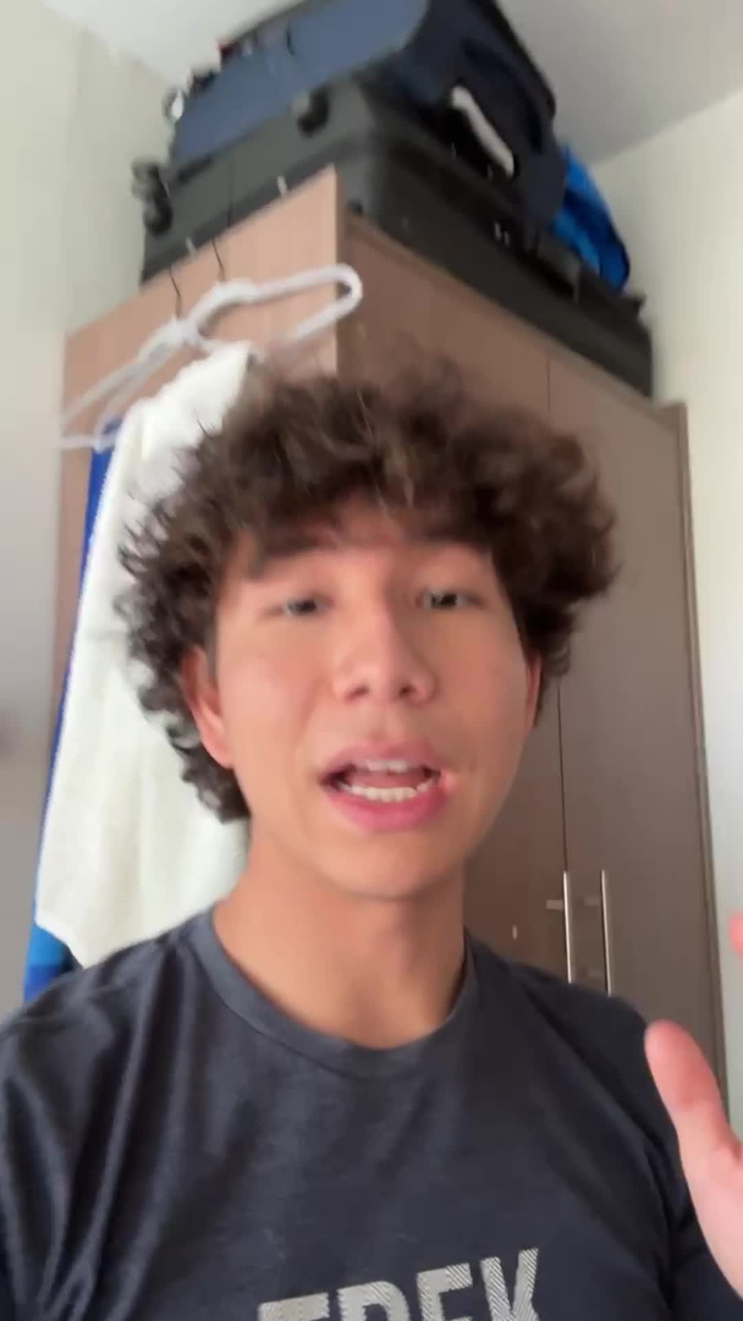 A young man with curly hair and a dark t-shirt is speaking directly to the camera. The shot is taken from a low angle, with the background being a room with a closet and luggage on top.