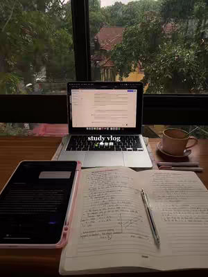 A well-lit image of a desk with a laptop, an open notebook, a tablet, and a cup of coffee is in front of a large window. The laptop screen displays a document, while the notebook has handwritten notes with a pen resting on it. A tablet lies to the left of the notebook and laptop. Through the window, the outline of the trees outside can be seen.