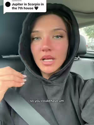 A woman with fair skin and light eyes in a dark hooded sweatshirt appears to be filming from inside a car. She is speaking with her mouth open, and the background shows a blurred view of the car's interior and a window.