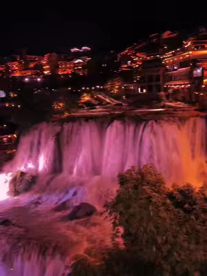 The cover image shows a nighttime landscape featuring a majestic waterfall as the focal point. The water cascades down, illuminated with shades of pink and purple lights, creating a vibrant contrast against the dark sky. At the top, a collection of buildings shines with warm orange and yellow lights, suggesting a town or city. The overall scene feels magical under the night sky.