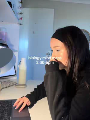 A young woman is seated at a desk, working on a laptop. The image is illuminated by soft, natural light from outside the frame, highlighting her face and the edge of the desk. Text overlays state 'biology mind maps 2:30-6pm'.