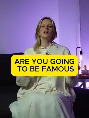 A woman sits in a relaxed pose in a well-lit room. A large yellow rectangle with black text reading "ARE YOU GOING TO BE FAMOUS" covers the lower chest and stomach area. The background fades to purple, and a small lamp and table are visible to the right. The woman is dressed in a light-colored, long-sleeved shirt, and has light blonde hair.
