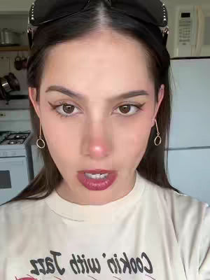 A close-up shot of a woman in a kitchen setting. She is the central focus, with dark sunglasses perched on her head. Below, she wears a white t-shirt that has red graphic print. Her skin is fair, with makeup highlighting her eyes and lips. The kitchen in the background is slightly blurred, showing elements like a stove, and appliances. The composition is well-lit, with the woman looking directly towards the viewer.