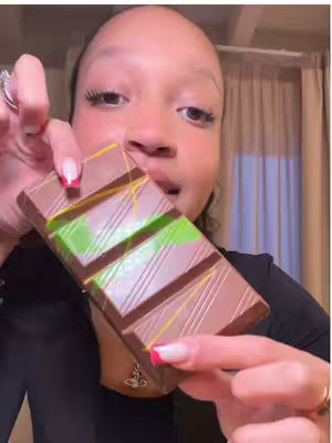 A medium shot of a woman holding a large chocolate bar. The woman is fair-skinned with dark hair and is looking towards the camera. She is wearing a black top and a finger ring with a large stone can be seen. She holds the chocolate bar near her face, with bright nail polish visible on her fingers. The chocolate bar is mostly brown with green stripes, possibly depicting a mint flavor. Behind the woman, a curtain gives a sense of background.