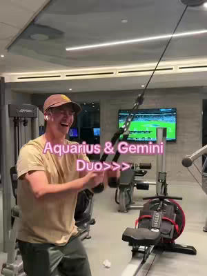 A man wearing a baseball cap and a light-colored t-shirt is working out in a gym. He's using a machine that appears to be for chest exercises.
