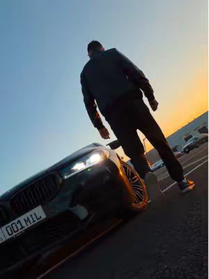 The cover image shows a man standing in front of a black car at a slight angle, with his back to the camera. The car is parked on asphalt, with some other cars and a building visible in the background under a gradient blue sky. The sky transitions into an orange hue near the horizon, suggesting a sunset or sunrise. The car's headlights are on, adding some visual interest. The man is wearing black pants and a jacket. The overall tone is stylish.