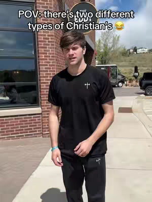 A young man is standing on a sidewalk in front of a brick building with a large window. He is wearing a black t-shirt and black pants. The image has a sunny, daytime feel.