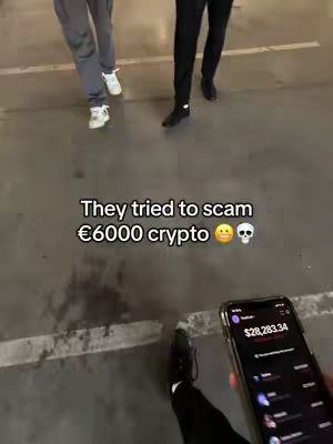 A first-person view, low-angle shot of a person holding a smartphone, likely recording a video. The phone screen displays financial data. The background features two people walking away from the camera. Text overlay: 'They tried to scam €6000 crypto' with emojis.