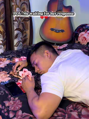 A man lies on a bed with a floral bedspread and a smartphone, with a guitar and a decorative headboard in the background. Text overlay states: POV: Me waiting for her response, with broken heart emojis.