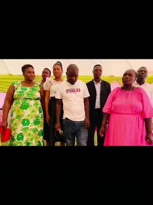 A group of people, consisting of 4 women and 4 men, are standing in front of a light pink and yellow backdrop. The person in the middle of the image, a man holding a microphone, is wearing a white t-shirt with a Polo logo. The women are in dresses and some are holding purses