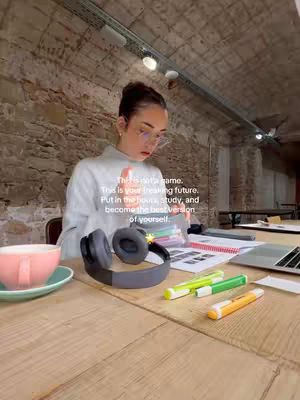 A young woman with her hair up in a bun is sitting at a light-wood table, immersed in studying with a pair of headphones and a cup. Text overlays read, 'This is not a game. This is your freaking future. Put in the hours, study, and become the best version of yourself.' The lighting is diffused.