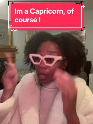 A woman with dark hair and pink glasses is in focus, with a red banner above her head. She is gesturing with both hands as if talking. The background is slightly blurred and contains some furniture.
