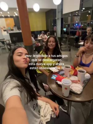 A selfie-style shot taken at a restaurant setting. Three young women are grouped around a small table. One in the foreground is taking the picture, while the two others are looking at the camera. The table has food items like burgers and drinks on it, suggesting a casual dining experience. The background shows the interior of the restaurant with other people, adding context to the setting.