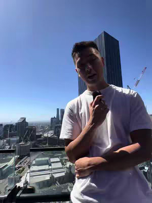 A person in a white t-shirt holds a black microphone against a cityscape backdrop and blue sky. The person is looking slightly down, with part of the hand covering the microphone.