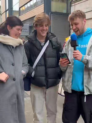 A candid street interview featuring three individuals. A woman in a gray coat stands to the left, a young man in a black puffer jacket and crossbody bag in the middle, and a man holding a microphone and phone on the right.