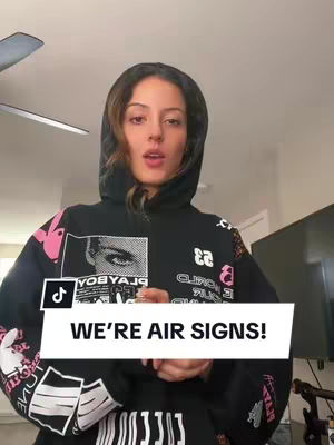 A woman with long brown hair wearing a black hoodie with a large printed design is centered in the frame. The hoodie is up, framing her face, and she has a neutral expression. Overlayed on the image is a white banner with black text that reads 'WE'RE AIR SIGNS!'