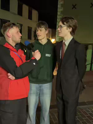 Three male figures are shown in an urban setting at night. One person, on the left, holds a microphone and is wearing a red vest over a dark jacket. The second person, in the middle, is wearing a green hoodie and light-blue jeans. The third person, to the right, is wearing a black suit. The background consists of buildings with some lights on.