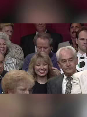A group of people sitting in an audience. The focus is on a woman with blonde hair, smiling and touching her hair. Other people are in the background, some smiling, creating a sense of engagement. The lighting is bright with primarily warm tones.