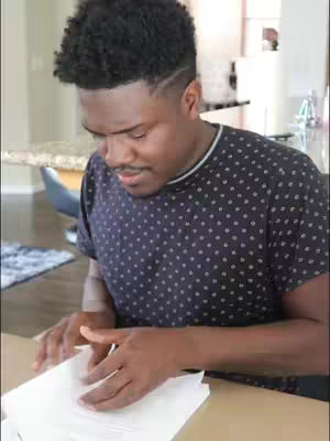 A man with dark skin and a short afro hairstyle looks down intently at a stack of papers. He wears a dark blue shirt with white polka dots. The background is a blurred interior space with a glimpse of a kitchen counter and a chair.