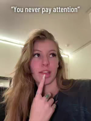 A woman with long blonde hair is looking up and to the right with a slightly surprised expression. She is pointing her index finger up towards her lips lightly, wearing a dark top and has multiple rings on her finger. Text above her says, "You never pay attention".