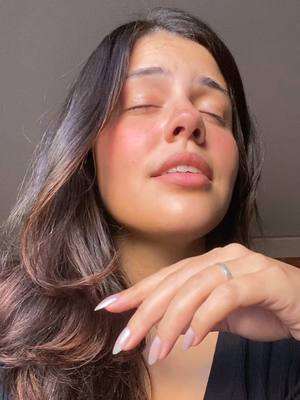 A close-up shot of a woman with long, dark hair and closed eyes. Her face is angled slightly upwards, and her expression suggests contemplation or emotion. She has a fair complexion with blush on her cheeks, and her lips are slightly parted. A slender silver ring is visible on her finger. The background is out of focus, with neutral colors.