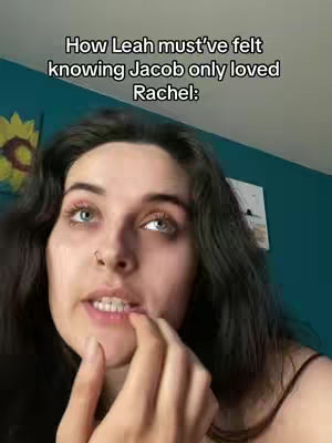 A young woman with long dark hair, a nose ring, and blue eyes gazes upwards, biting her lower lip with her index finger. The composition is framed closely, focusing on her face, with a slightly blurred turquoise wall and a partial sunflower image in the background.