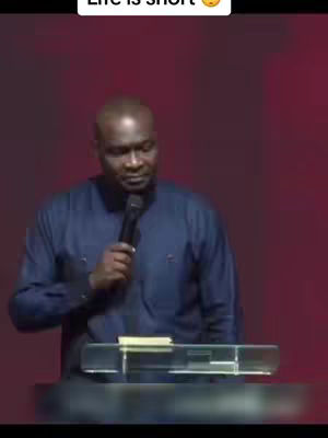 A man in a dark blue shirt stands behind a clear podium, holding a microphone. The background is a dark red.