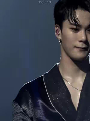 A medium shot of a man, likely in his 20s or 30s, with dark hair slicked back slightly. He has a fair complexion and a subtle smile. He is wearing a dark, possibly satin-like, blazer with a contrasting white trim and a silver chain necklace. The background is a deep blue, creating a contrast with the man's features and attire.