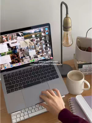A person's hand rests on a laptop trackpad, with a cluttered screen visible. Beside the laptop is a desk lamp with a large bulb, a mug of coffee, and a stack of books. The setup suggests a workspace.