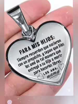 A close-up shot of a silver heart-shaped pendant held in a light-skinned hand. The pendant has a silver heart pendant design with text in Spanish.
