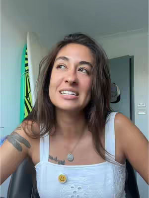 A young woman with long brown hair is looking up and to the side, smiling slightly. She's wearing a white tank top with a button that says "Smile". She has a tattoo on her upper arm and a small tattoo on her chest. Behind her is a surfboard, and a gray door.