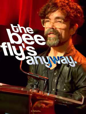A man with short, curly brown hair, glasses, and a beard stands at a podium, speaking into a microphone. The background is a blurred red, and the foreground features a black podium and a microphone. Superimposed over the image is the text, 'the bee fly's anyway.'
