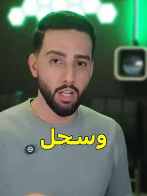 A man with a beard and a serious expression is centered in the frame. He wears a gray t-shirt. The top and right sides of the background include green lighting, indicating an indoor setting. Text overlay in Arabic is visible.
