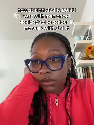 A woman with dark skin and braided hair is situated indoors, gazing slightly downward with a thoughtful expression. She wears large, square blue glasses and a red hooded sweatshirt. Text above her head reads "how straight to the point I was with men once I decided to be serious in my walk with Christ."