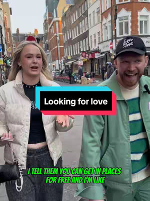 A casual shot of a man and woman walking down a city street. The woman is on the left looking at the camera with a neutral expression and open mouth. The man is on the right, also looking at the camera and smiling. Overlaid on the image is a black bar with white text that reads "Looking for love" and another text overlay that says "I TELL THEM YOU CAN GET IN PLACES FOR FREE AND I'M LIKE".