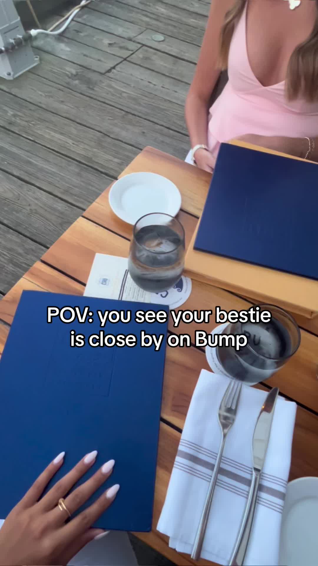 An overhead shot captures a table set for a meal on a wooden deck, with a woman's hand and manicured nails prominently in view. A blue menu or document, a partially visible white plate, and a tall glass with a drink and ice are placed on the table. The setting suggests an outdoor dining experience, possibly at a restaurant. The overall composition is well-lit, showing a casual dining setting, and the text overlay provides context that the bestie is near by. This composition sets a friendly or social tone inviting viewers into a personal experience.